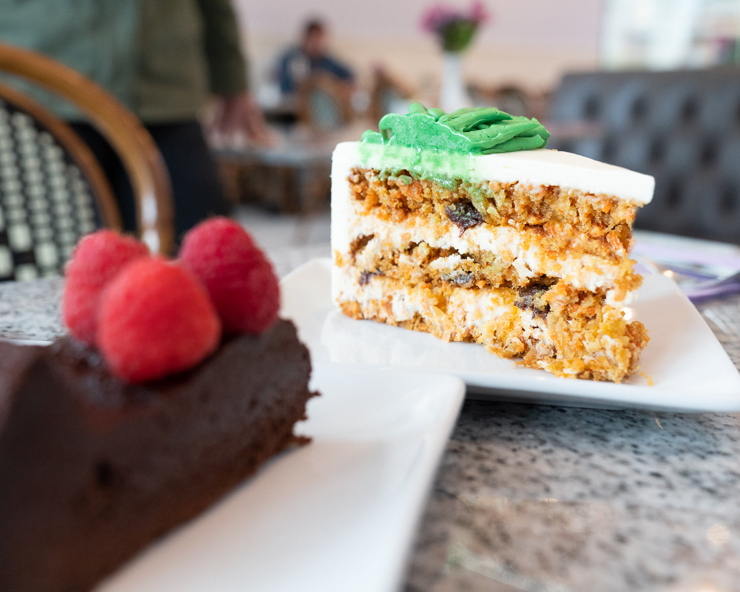 Slice of carrot cake with green frosting on a plate, surrounded by other cakes on a table.