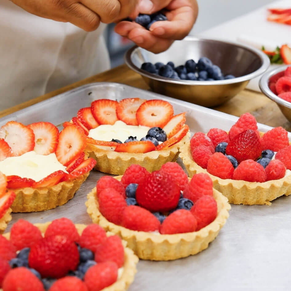 A baker carefully placing fresh blueberries and strawberries onto fruit tarts in a dedicated gluten-free kitchen.